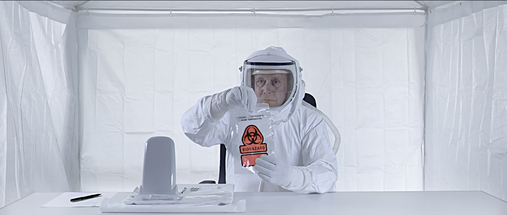 Man in spacesuit holds a bag labelled "Biohazard" in a square white tent-like environment.