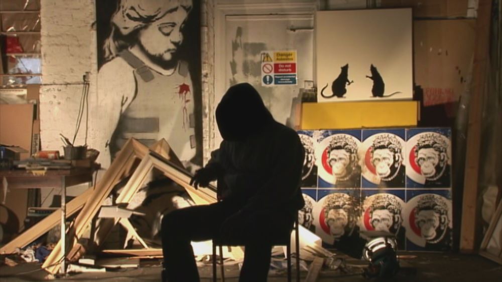 A person in a hooded jacket and sitting on a chair is silhouetted in what appears to be a painting workshop, surrounded by images of rats, ape faces, and a girl with a bloodied spot on her chest.