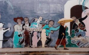 Older woman with umbrella and a cane walks past a colorful outdoor mural.