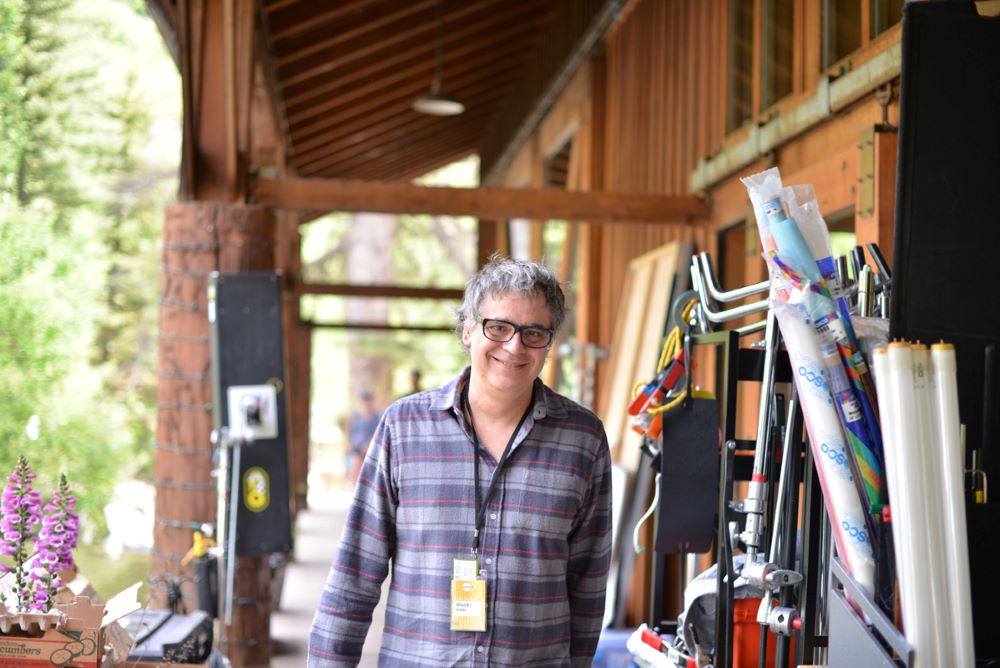 Gray-haired, bespectacled man in purple plaid shirt and lanyard smiling at the camera and walking on the boardwalk or porch at the front of a wooden building, with what appears to be umbrellas, outdoor chairs, and other equipment are stored