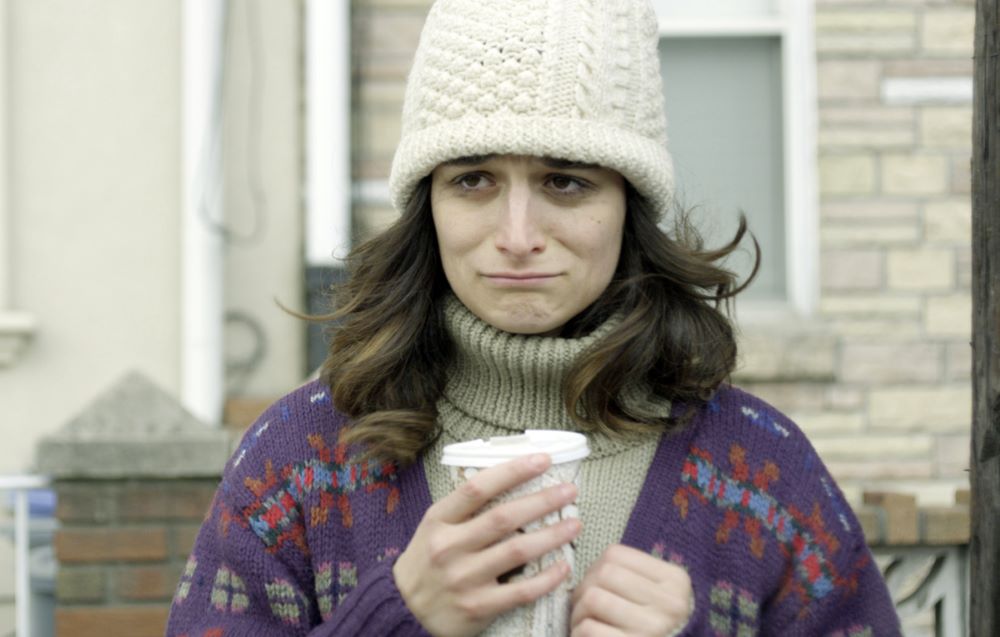 Young woman with long brown hair, wearing beige stocking cap, purple print cardigan over darker beige turtleneck sweater and holding what appears to be a disposable cup with a lid, standing in front of a building. She has a comically pouty expression.