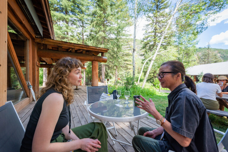A woman with light brown curly hair wearing a dark sleeveless top talks to a woman with black hair tied back in a blue shirt and glasses. Both are sitting in front of a cabin at the Sundance Mountain Resort.