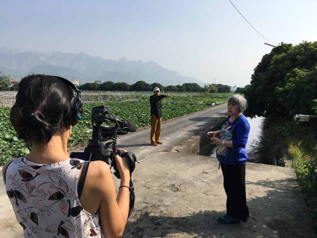 Image foreground shows the filmmaker’s back, holding a camera, with a person in a bluish purple shirt with a bridge design toward the right side of the frame. Green crops extend along the left of the image, with a road cutting diagonally upward from left to right. Standing on the road, another person is gesturing toward the right side of the frame.