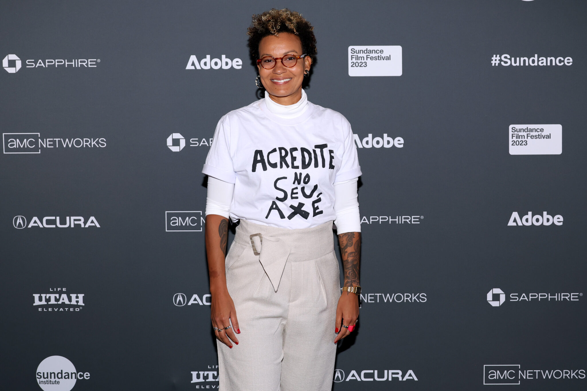 A Black woman with short hair wearing glasses, a white long sleeve, and tan pants, stands in front of a step-and-repeat at the 2023 Sundance Film Festival.