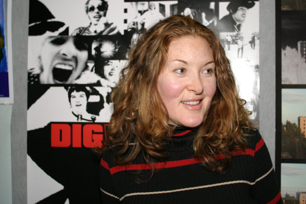 A close up of woman with medium-length, red, wavy hair in a black sweater with red stripes as she smiles on. A poster of the movie "DIG" is behind her.
