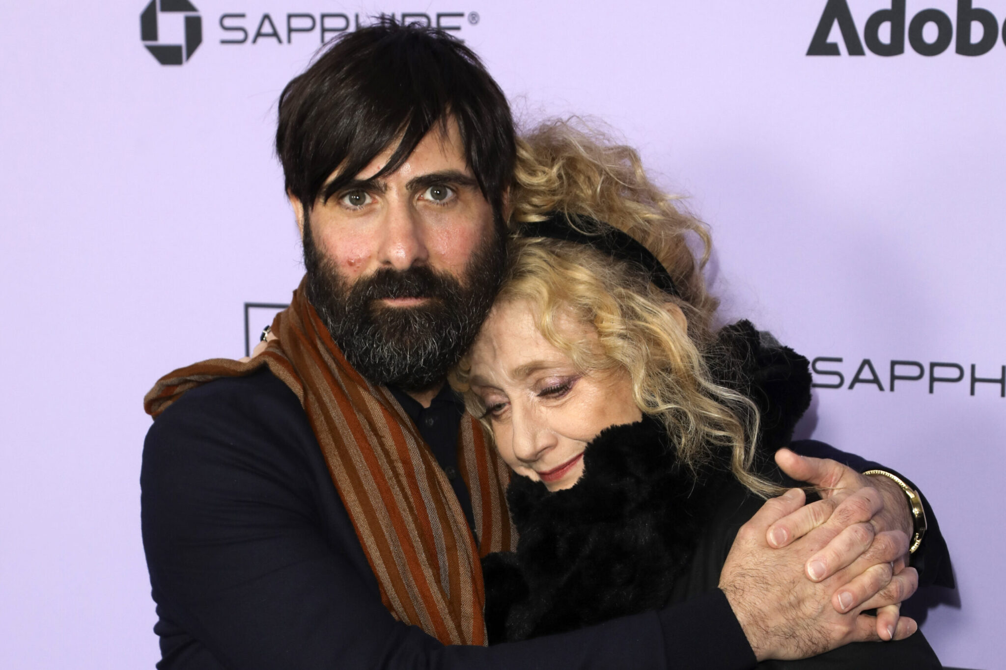 A white man with a beard and dark brown hair hugs an older white woman with curly blond hair in front of a purple backdrop