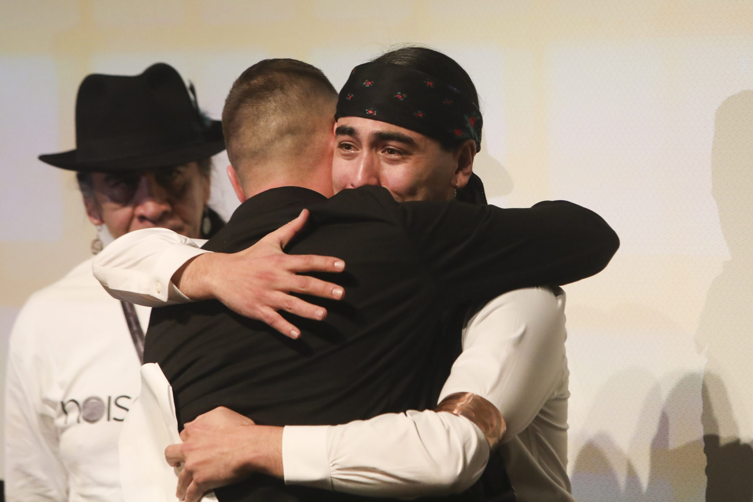 Indigenous man, tears in his eyes, hugs a man in a dark suit jacket on what appears to be a stage. Another man watches in the background.