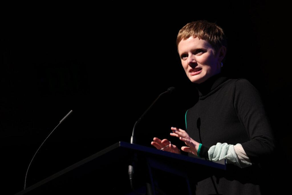 Rose Glass stands at a podium and speaks into a microphone against a black backdrop.