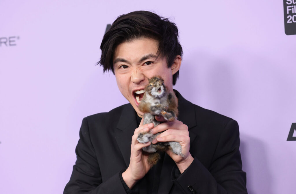 Posing in front of a 2024 Sundance Film Festival backdrop, Sean Wang shows off a squirrel toy to the camera.
