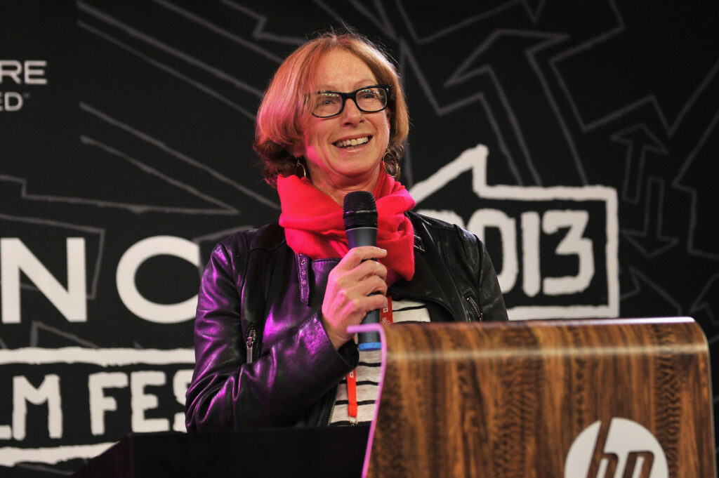 A woman with short red hair and glasses in a red scarf and black leather jacket, stands at a podium and speaks into a mic.