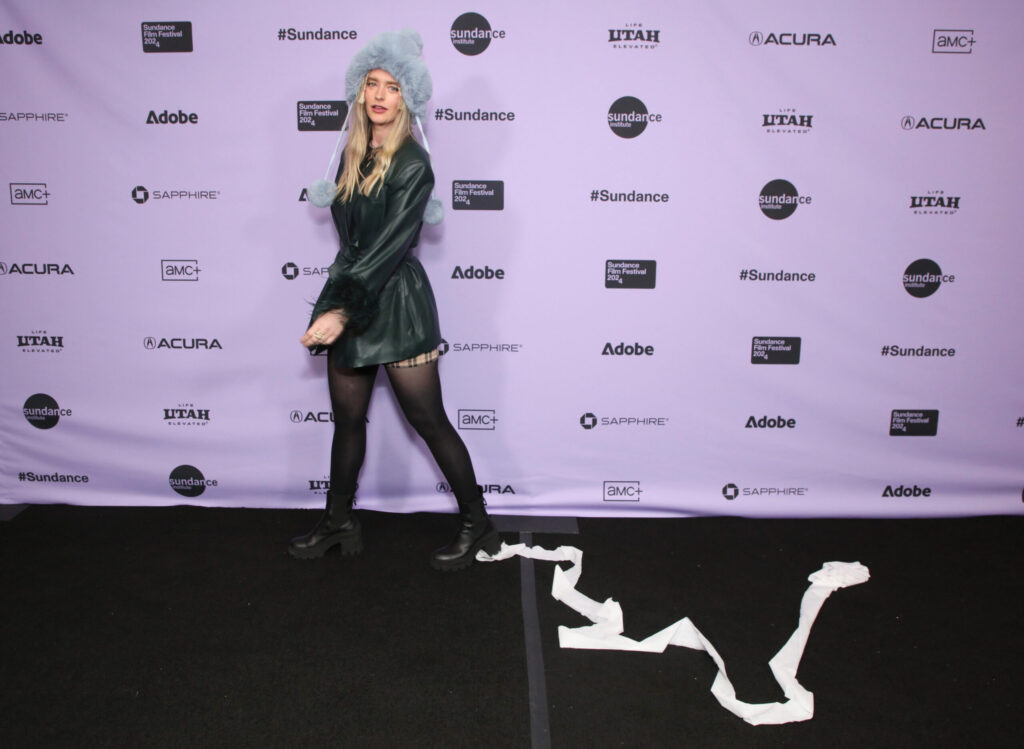 a blonde woman with a furry hat stands in front a a purple step-and-repeat with toilet paper hanging from her shoe.