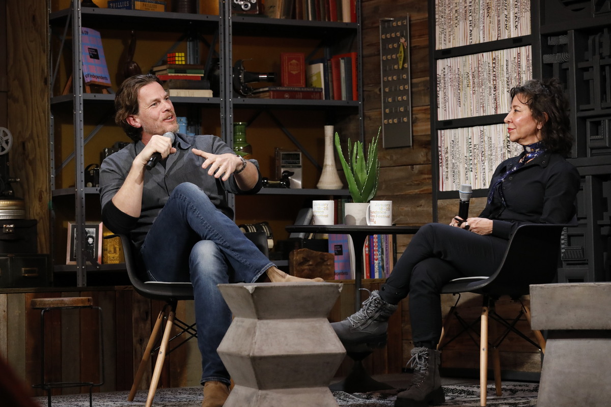 A white man with brown hair sits down and speaks into a microphone next to a woman with dark hair wearing a black sweater