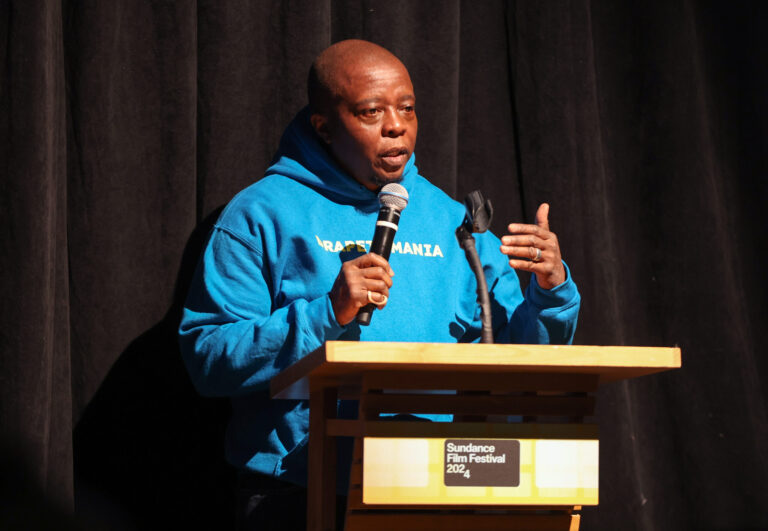 Director Yance Ford stands at a podium holding a microphone. He is wearing a blue sweatshirt.