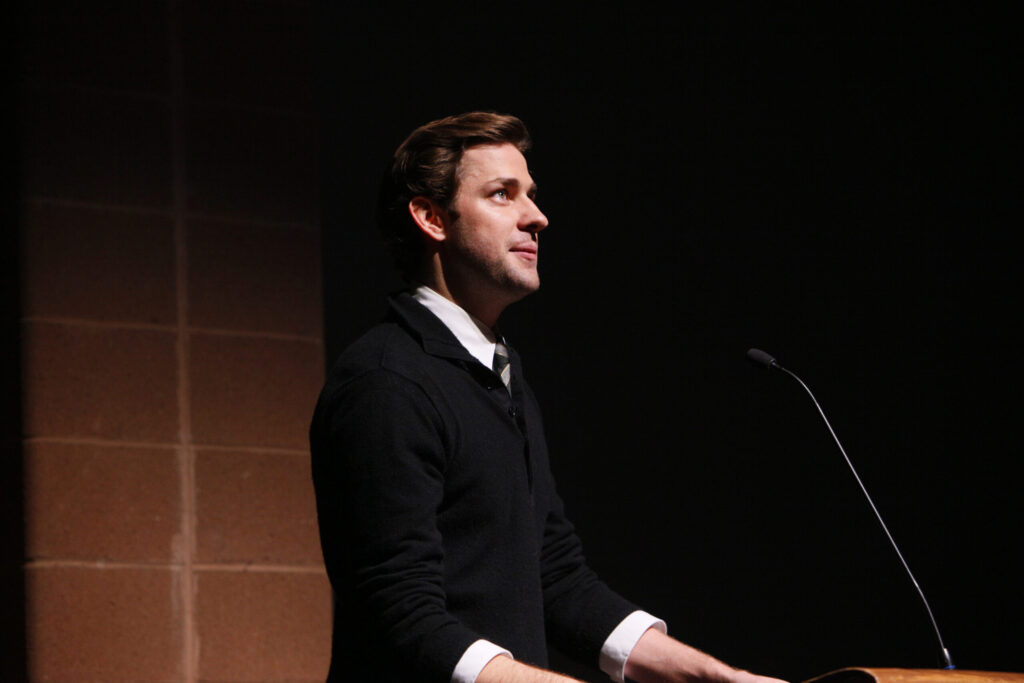 A man with brown hair in a black sweater with a white collar stands in a theater and looks ahead at the audience