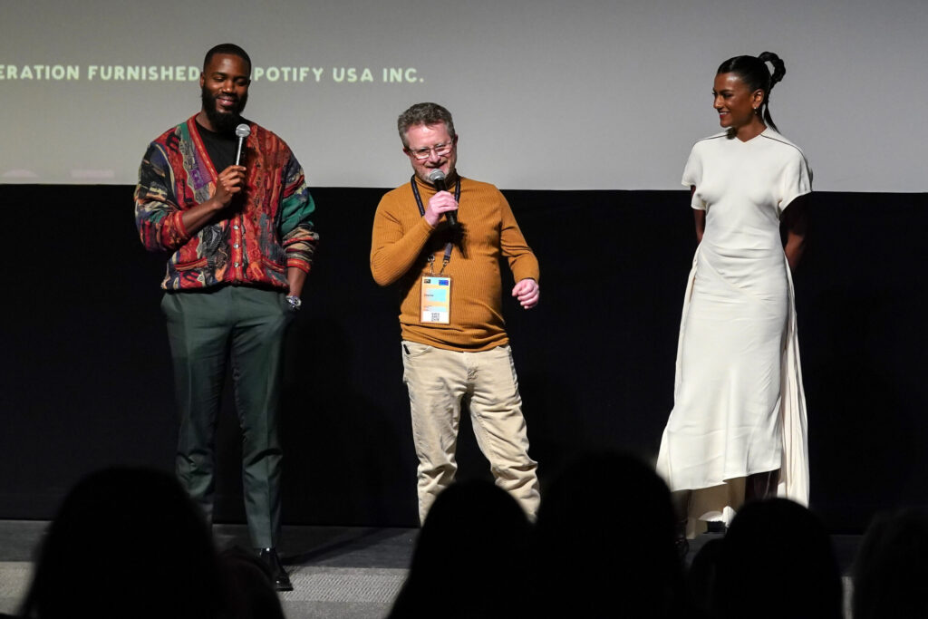 A black man in a colorful sweater holding a mic, a white man in an orange sweater holds the mic, and a woman in a white dress, stand on stage speaking to audience members