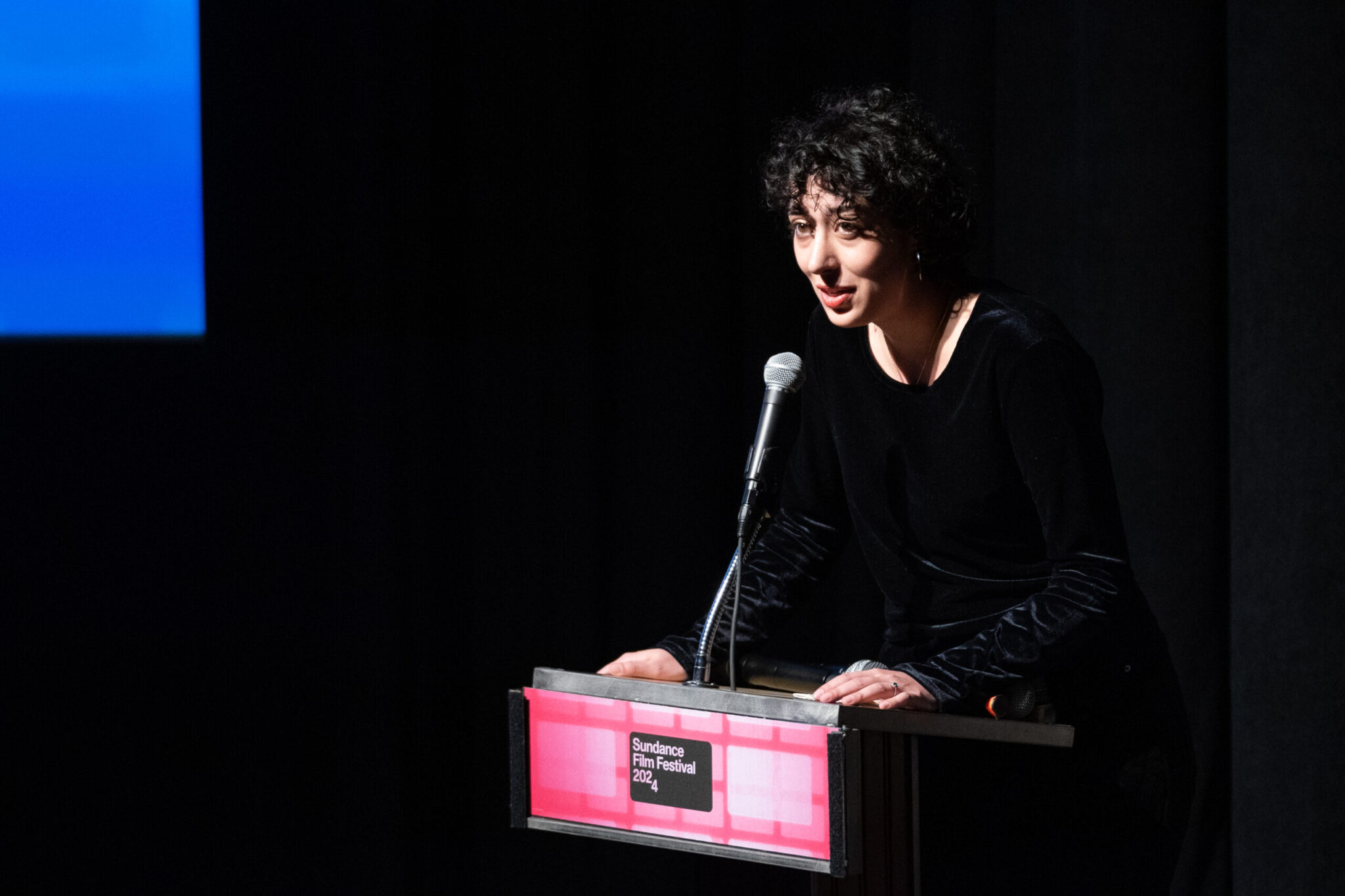 Woman with short, curly dark hair and dressed in black, steps up to a podium at the 2024 Sundance Film Festival.