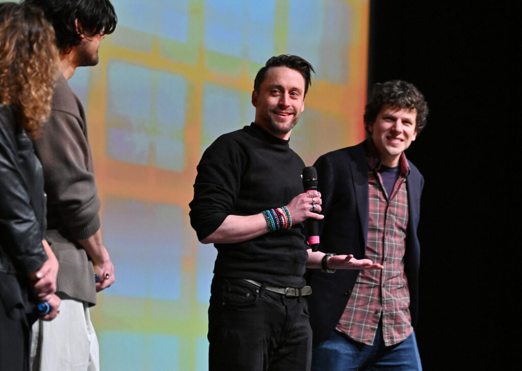 Kieran Culkin holds a microphone in front of a multicolor 2024 Sundance Film Festival backdrop. Jesse Eisenberg stands to his right.