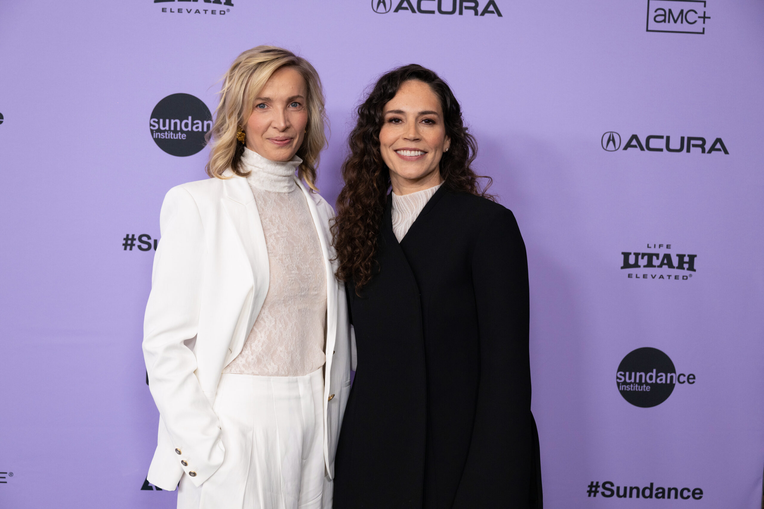 A blonde woman in a white pant suit stands next to a woman with long curly brown hair in a black coat