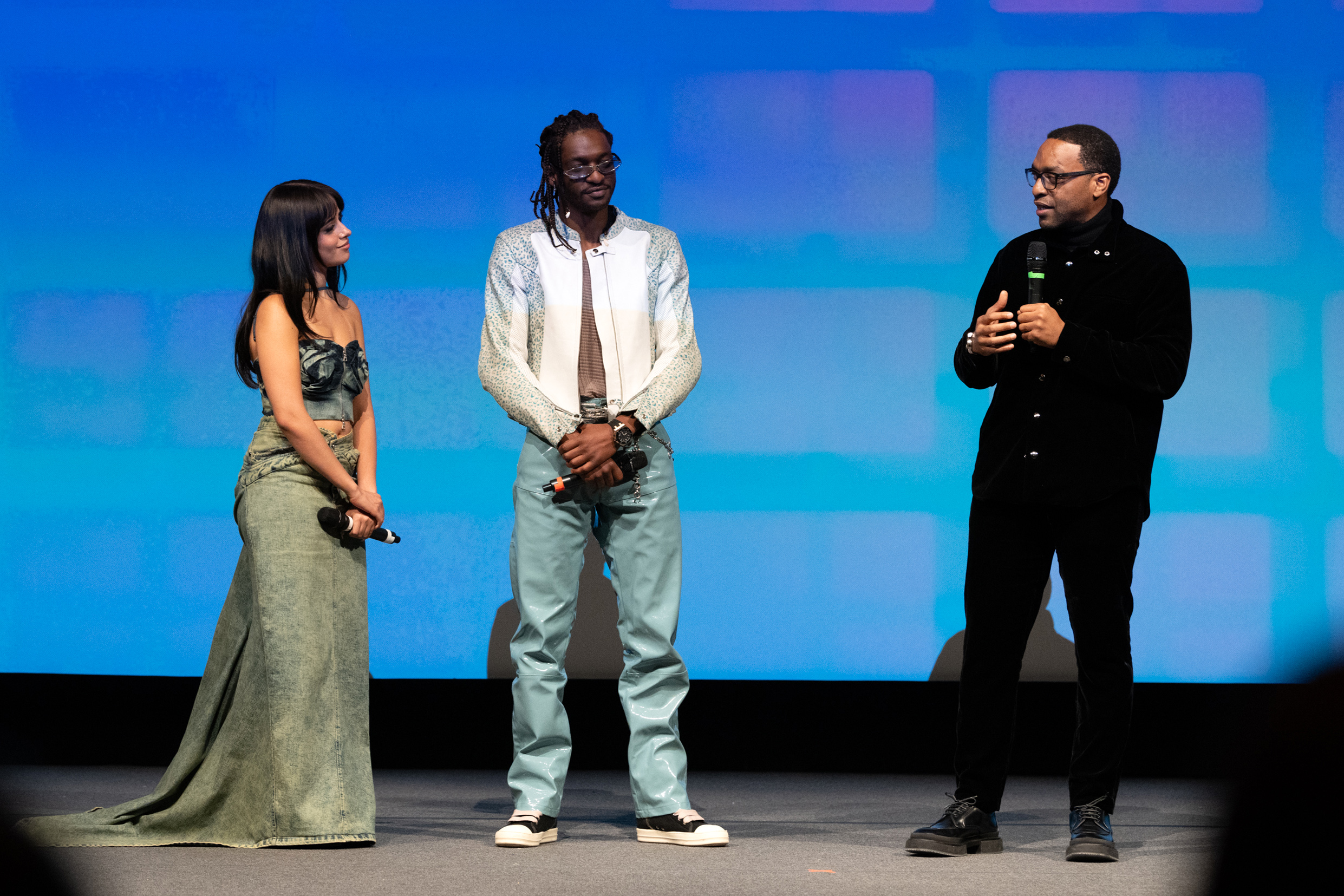 A Mexican-Cuban woman with long black hair in a denim outfit, a Black man with a white dress shirt and blue pants, and a Black man dressed in all black, stand on stage at Eccles Theatre.