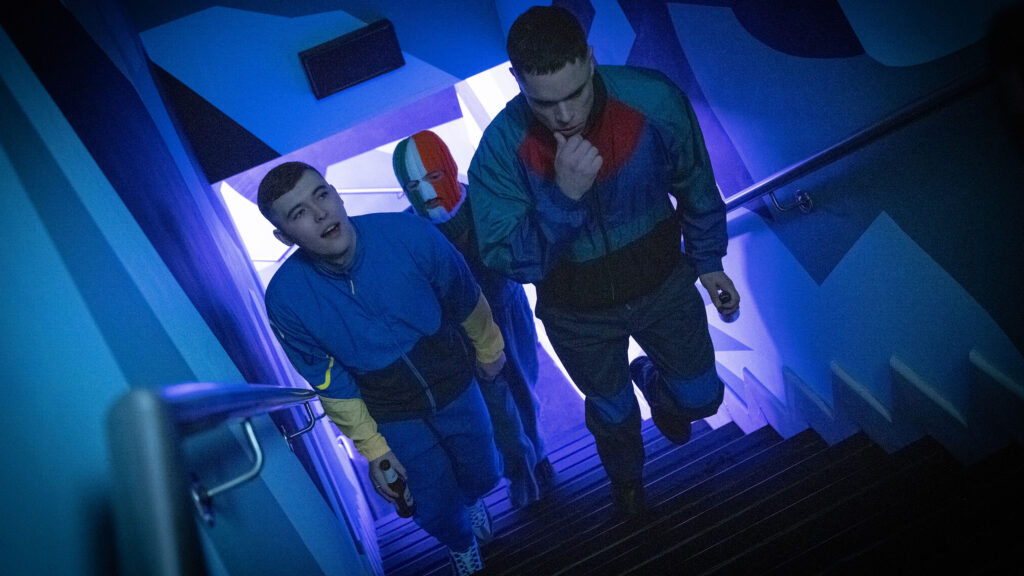 A film still from Kneecap. The three members of hip-hop trio Kneecap walk up the stairs in blue-tinted lighting.
