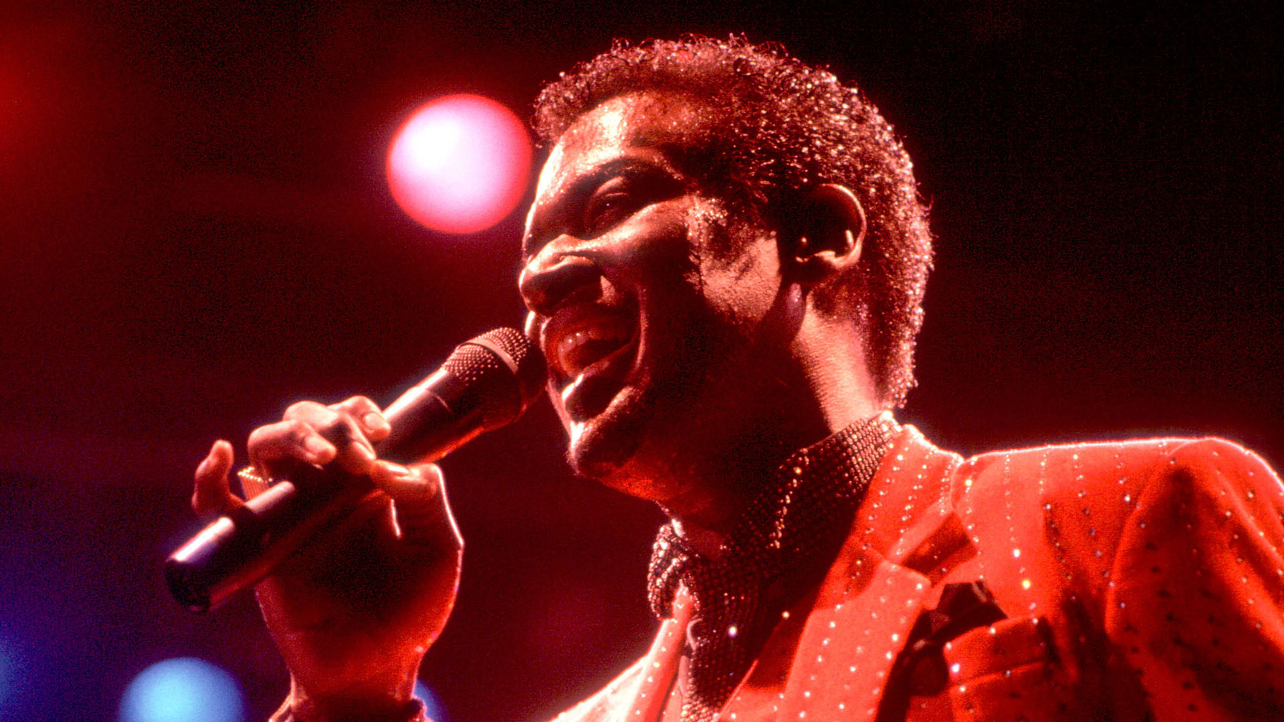 A film still from Luther: Never Too Much that shows Luther Vandross singing into a microphone in red-tinted lighting.