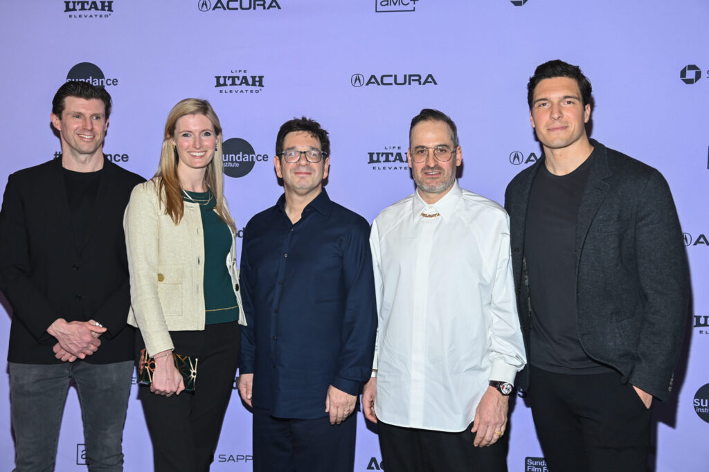 A man with brown hair in a black coat and jeans, a blonde woman with a green blouse, white jacket, and black pants, a man with brown hair and glasses, a blue dress shirt, and black pants, a man with brown hair and glasses, a white dress shirt and black pants, and a man with black hair , a black jacket, and black pants, all stand in front of the 2024 Sundance Film Festival step-and-repeat.