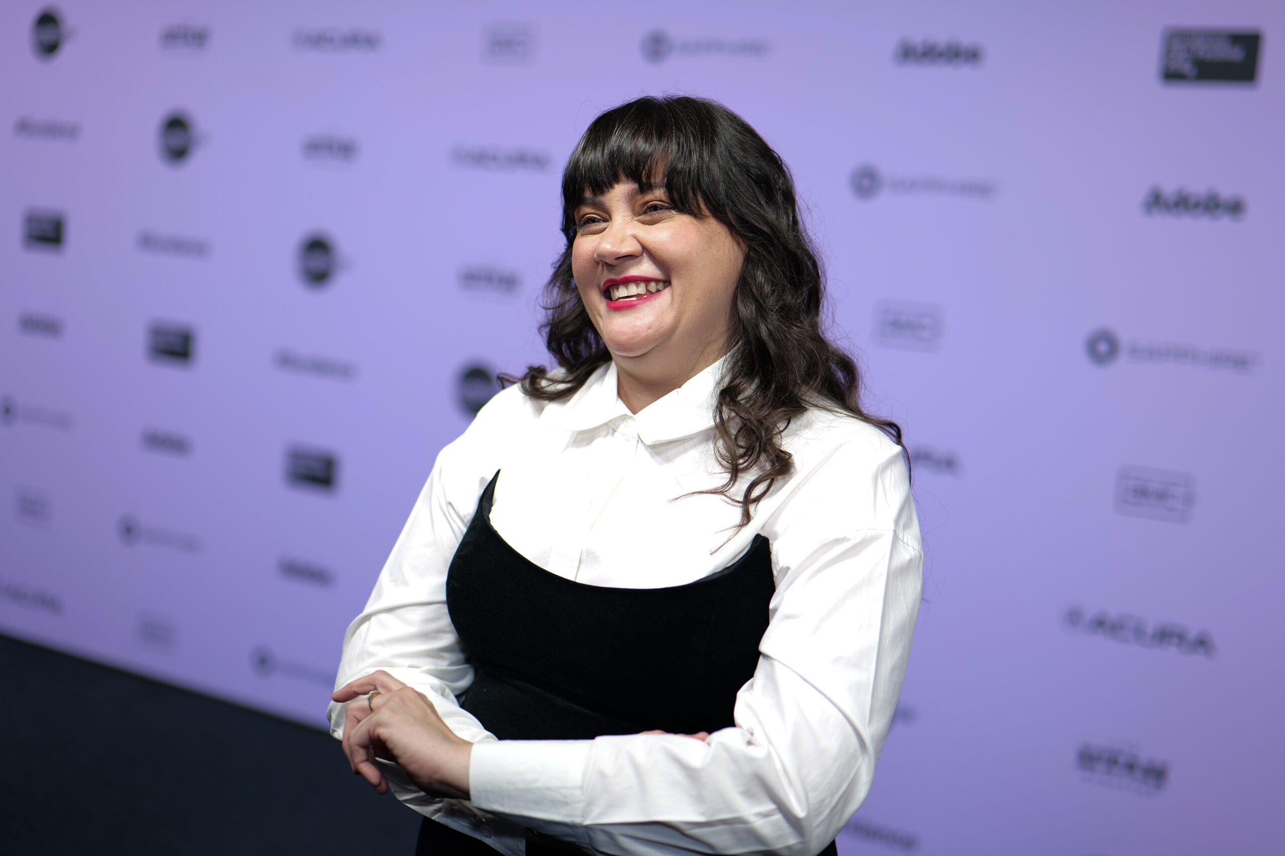 A woman in a long sleeve white tee with a black dress over, stands in front of a step and repeat.