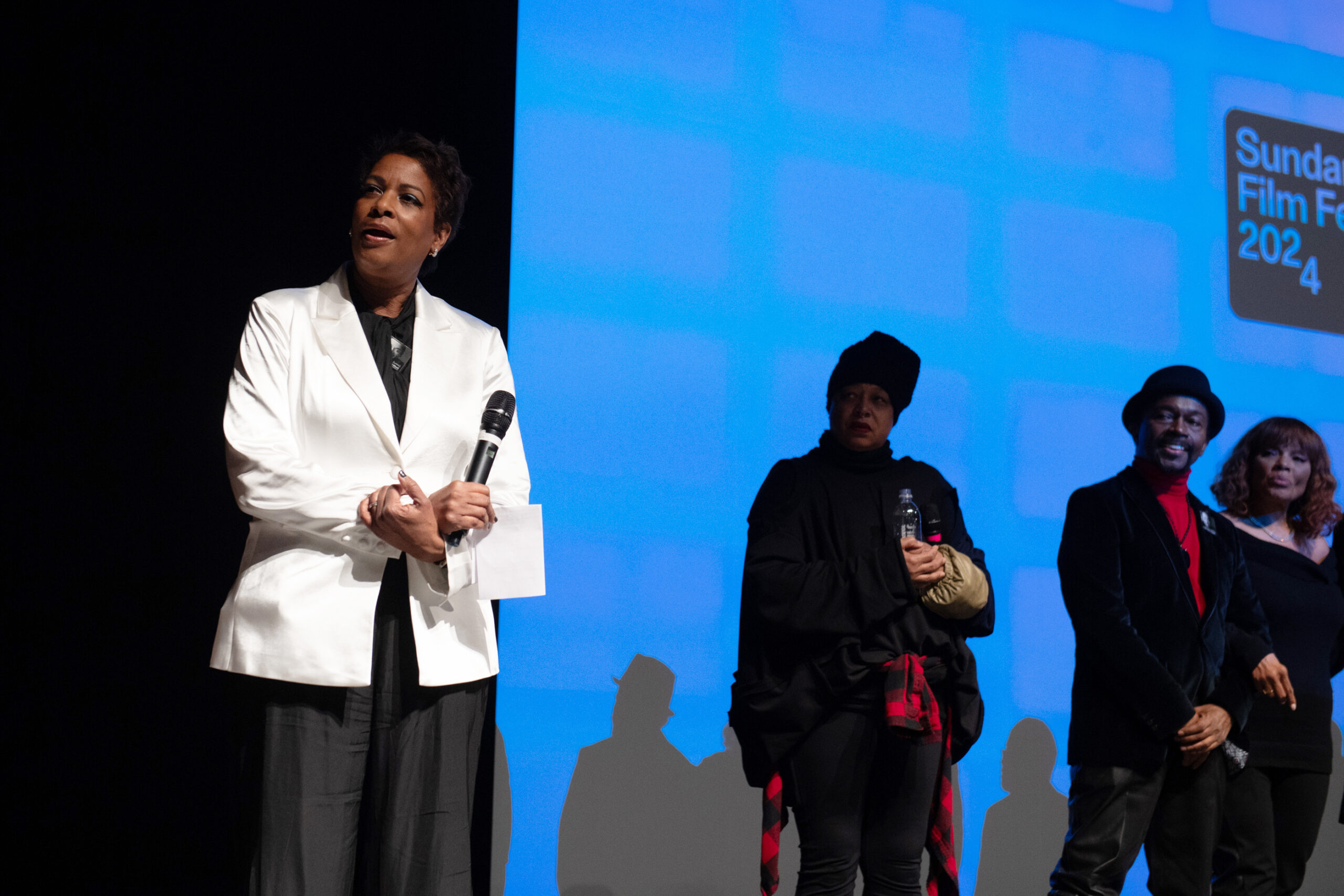 A Black woman with short hair in a white blazer holds a mic on stage. The Sundance Film Festival 2024 logo is on the screen behind her.
