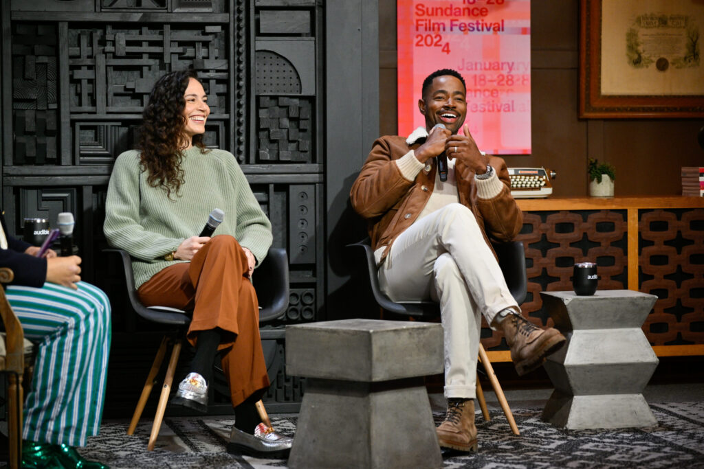 A woman with long curly hair in a green sweatshirt and brown pants sits next to a Black man with white pants and a tan jacket.