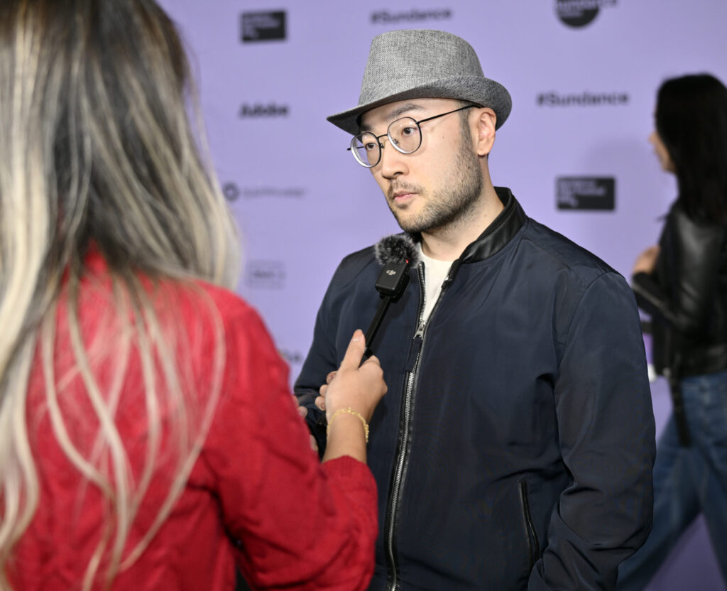 Asian man with short beard and glasses, wearing a gray fedora, is being interviewed