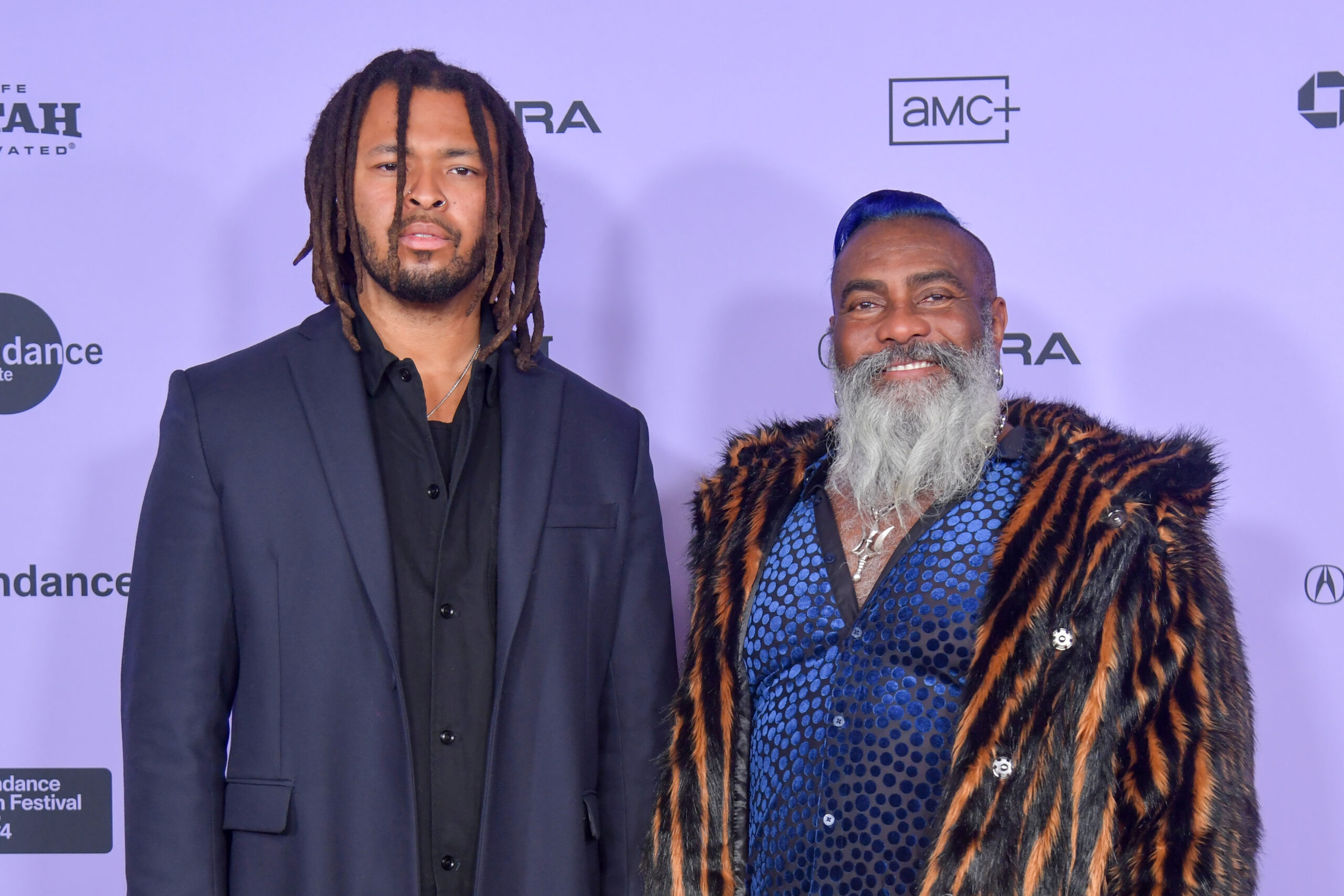 Two Black men, one with a beard and dreadlocks, the other with a long gray beard and wearing an orange-and-black-striped coat, stand in front of a Sundance Film Festival backdrop.