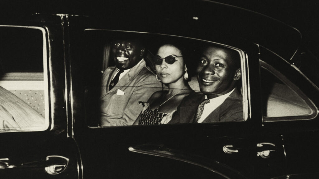 A film still from Soundtrack to a Coup d'Etat. The black and white image shows Andrée Blouin and Patrice Lumumba sitting in the backseat of a car.
