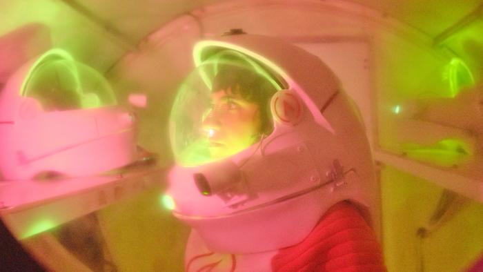 A woman with black hair in a space helmet stares at another space helmet with her eyes wide open