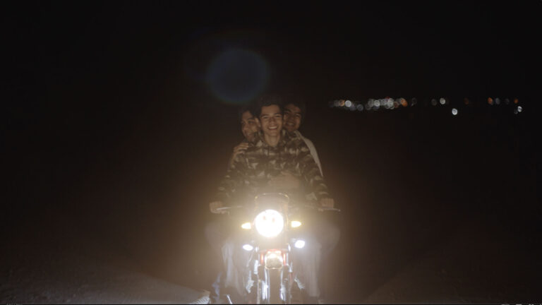 A young teen rides a motorcycle with two others riding on the back. City lights are in the background.