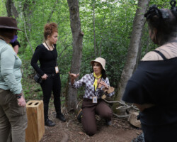 Lab fellow Caledonia Curry (center, left) and Laura Merians Gonçalves (center, right) prepare the set for Caledonia's project 'Sibylant Sisters.' Photographer: Ash Gerlach Production crew in conversation in a green woodsy area