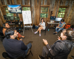 Lab fellows and creative advisors convene to discuss 'Standing Above the Clouds.' Photographer: Miguel Mendoza Individuals sit in a circle near a whiteboard