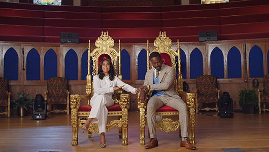 A dressed-up Black couple sit in side-by-side gold and red velvet thrones