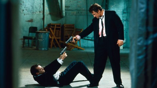 A man in a suit, with what appears to be blood on his shirt, points a semi-automatic pistol and a man lying on his back on the floor, who points a semi-automatic pistol at the standing man. They appear to be inside a warehouse type of building.